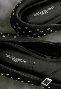 Scarpe nere con una tomaia testurizzata decorata con borchie argentate, una finitura simile alla seta e una cinghia con una fibbia decorativa. Etichette visibili all'interno.
