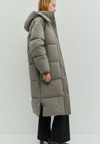 Lange grijze donsjas met capuchon, voorzien van horizontale doorgestikte secties, knopen aan de voorkant en zijopeningen voor bewegingsvrijheid.