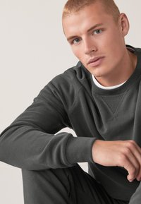 Tumehall sweatshirt, millel on ümar kaelus, tekstureeritud kanga ja nähtavate õmblusdetailidega. Mudelil on heledat nahka ja lühikesed juuksed.