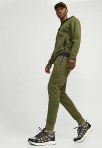 Olive green tracksuit with black accents, featuring zippered pockets, and paired with black and grey sneakers. Smooth, elastic material.