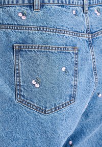 Denim kratke hlače z zadnjim žepom, okrašenim z vezenjem rožnatega cveta češnje in rahlo teksturiranim modrim pranjem. Opažene so kontrastne šive.