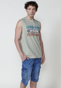 Bez piedurknēm gaiši zaļa krekls ar grafisku druku, kombinēts ar ziliem cargo šortiem, kuriem ir vairāki kabatas un brīvs piegriezums.