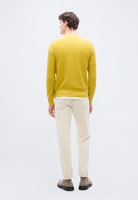 Homme portant un pull moutarde texturé, un pantalon beige clair et des bottes en daim grises, debout de dos sur un fond blanc uni.