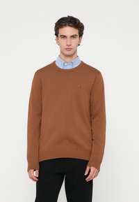 Καφέ πλεκτή ζακέτα με στρογγυλή λαιμόκοψη, με υφασμάτινη υφή και ριμπ μανσέτες, φορεμένη πάνω από ένα ανοιχτό μπλε πουκάμισο με γιακά, συνδυασμένη με μαύρο παντελόνι.