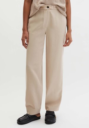 Pantaloni beige a gamba larga con bottone e zip, abbinati a mocassini neri slip-on e una maglia beige strutturata infilata dentro.