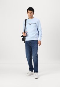 Šviesiai mėlynas apvalios iškirptės megztinis su "TOMMY HILFIGER" logotipu, derinamas su tamsiai mėlynomis džinsais ir baltais kedais. Modelis laiko juodą petinę rankinę.