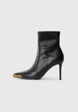 Black leather ankle boot with a sleek, fitted design. Features a pointed toe with a gold metal accent and a high stiletto heel.
