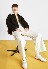 Hnědá, oversized, strukturovaná bunda se světle zeleným límcem, nošená přes béžový top. Bílé široké kalhoty a tenisky s růžovými akcenty.
