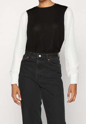 Persoon draagt een zwarte mouwloze top over een witte blouse met lange mouwen met structuur, gecombineerd met zwarte high-waisted jeans.