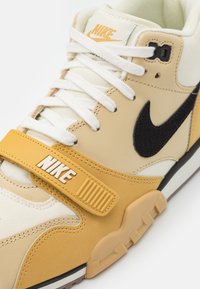 Béžové a žluté tenisky s texturovaným svrškem, černým logem Nike, bílými tkaničkami a páskem s logem Nike. Gumová podrážka.