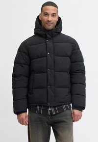 Chaqueta de abrigo negra con capucha, que presenta paneles acolchados, botones a presión, dos bolsillos y puños de canalé. Llevada sobre una camisa de cuadros.