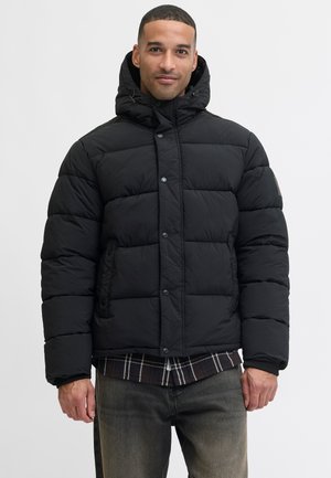 Chaqueta de abrigo negra con capucha, que presenta paneles acolchados, botones a presión, dos bolsillos y puños de canalé. Llevada sobre una camisa de cuadros.
