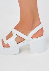 Sandali bianchi con tacco, dotati di cinturini incrociati regolabili, realizzati in materiale lucido. Tacco quadrato spesso e design con punta aperta. Texture liscia.