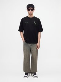 Νεαρός άνδρας που φοράει μαύρο γραφικό T-shirt, γκρι παντελόνι με φαρδιά πόδια, ασπρόμαυρα αθλητικά παπούτσια και σκούρα γυαλιά ηλίου, στέκεται μπροστά σε λευκό φόντο.