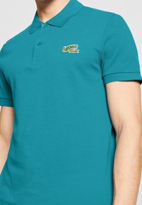 Turkos pikétröja, bomullstyg, korta ärmar, ribbad krage, har en liten broderad Lacoste-logotyp på vänster bröst.