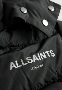 Čierna nylonová bunda s striebornými cvokmi a výrazným logom "ALLSAINTS LONDON" v tučnom bielom písmene. Hladký povrch.