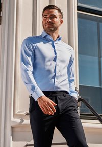 Lichtblauw button-down shirt met een gladde textuur en een gestructureerde kraag, gecombineerd met donkere broeken, tegen een moderne architecturale achtergrond.