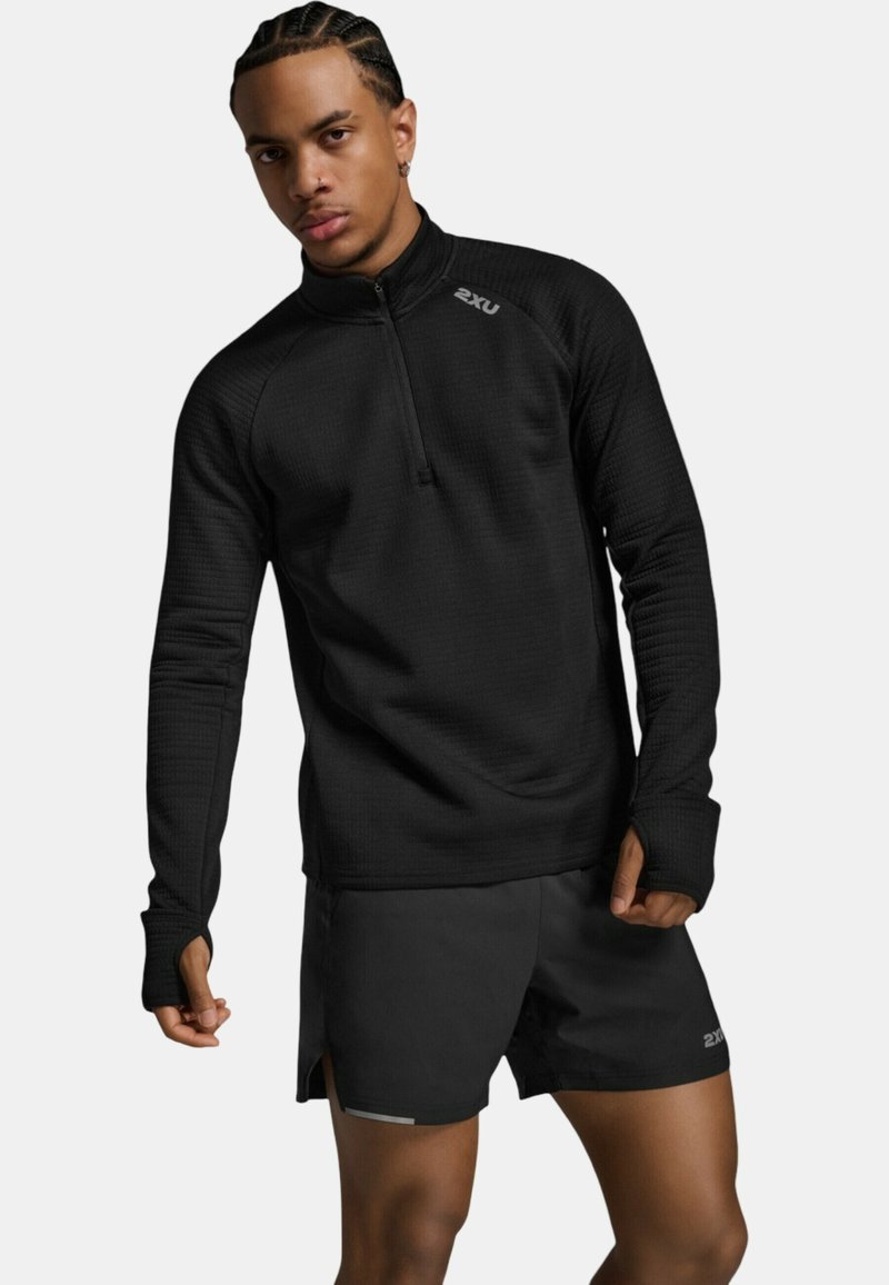 Mand med flettet hår iført sort langærmet atletisk trøje og sorte sportsshorts, poserende mod en ensfarvet baggrund.