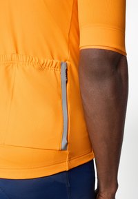 T-shirt de sport orange à manches courtes, avec un ourlet froncé et un détail de poche réfléchissante grise à l'arrière. Texture de tissu lisse.