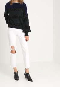 Musta ja laivastonsiniraidallinen neule teksturoiduilla yksityiskohdilla, yhdistetty kuluneisiin valkoisiin farkkuihin, jotka ovat polvissa revityt, sekä mustiin nilkkureihin, joissa on korko.