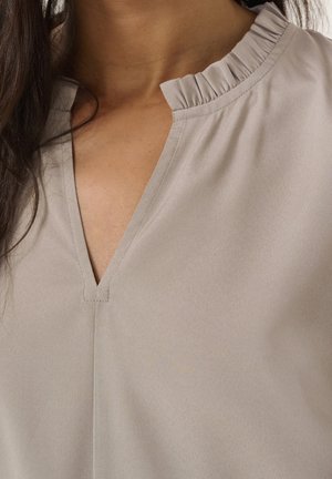 Femme portant un chemisier beige avec un petit col à volants et un décolleté en V, laissant apparaître le cou et le haut de la poitrine.