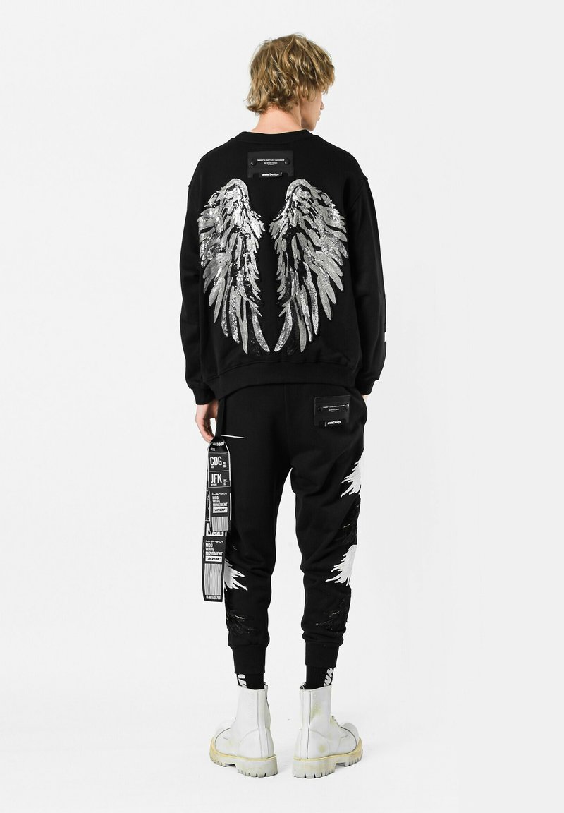 Sudadera negra con gráficos de alas plateadas en la parte posterior, combinada con pantalones negros que tienen acentos blancos y varios parches gráficos. Botas blancas.