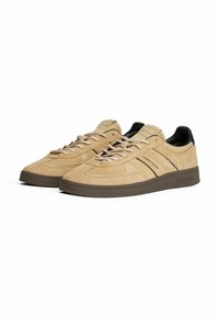 Beige Wildleder Sneakers in Low-Top-Form mit braunen Akzenten, versehen mit Schnürverschluss, minimalem Branding an den Seiten und einer strukturierten braunen Gummisohle.