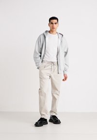 Grå sweatshirt med hætte, hvid t-shirt, beige bukser med snor, og sorte sko. Enkelt, afslappet design med løs pasform og ensfarvede nuancer.
