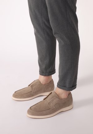 Beige suede slip-on shoes with white rubber soles, featuring stitching details and a smooth finish, paired with dark gray rolled-up trousers.