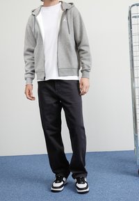 Sudadera gris con cremallera y detalles en blanco, llevada sobre una camiseta blanca, combinada con pantalones negros y zapatillas deportivas en blanco y negro. Diseño sencillo.