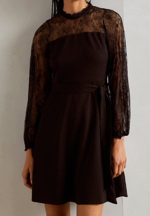 Vrouw die een zwarte jurk draagt met doorschijnende kanten lange mouwen, een hoge kanten halslijn en een tailleband met strik, tegen een effen achtergrond.