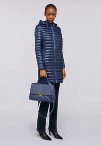 Giacca puffer blu navy con zip frontale e cappuccio, abbinata a jeans in denim scuro e décolleté nere a punta, mentre tiene una borsa strutturata.