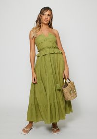 Woman wearing a sleeveless olive green tiered maxi dress and white sandals, holding a woven straw handbag with gold handles.