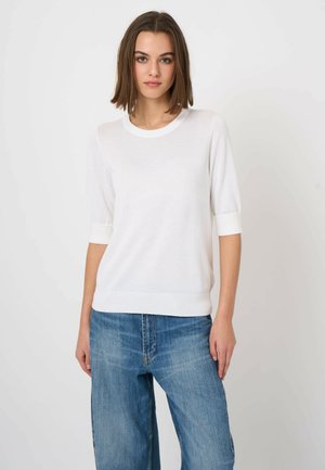 Young woman with shoulder-length brown hair wearing a white short-sleeve sweater and blue denim jeans, standing against a plain background.