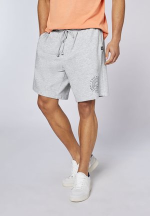 Mann trägt hellgraue Shorts mit Kordelzug und Logo am linken Bein, pfirsichfarbenes Shirt und weiße Sneakers, steht vor einfarbigem Hintergrund.