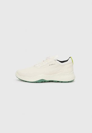 White athletic shoes with a textured cream upper, green rubber sole, and contrasting black inner lining. Features laces and a neon accent.
