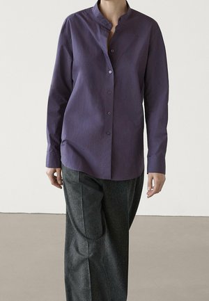 Personne portant une chemise violette à manches longues boutonnée avec un col mao et un pantalon large gris sur un fond uni.