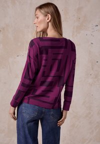 Top morado de manga larga con patrón geométrico rosa, con un escote ancho y un corte relajado, combinado con jeans de mezclilla azules.