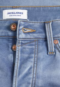 Jeans de mezclilla azul claro con una etiqueta cosida, herrajes de cobre, cierre de botón y costuras amarillas doradas en contraste.
