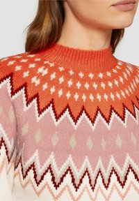 Gebreide trui met een hoge hals, grote zigzagpatronen in oranje, roze en roomkleur, met kleine witte geometrische vormen die door de stof heen zijn verwerkt.