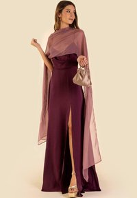 Abito da sera bordeaux con spacco alto sulla coscia, accompagnato da una delicata sciarpa color malva e una borsa dorata chiara. Presenta un design aderente e tacchi a fascia.