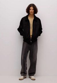 Giubbotto bomber nero con zip davanti, indossato sopra una t-shirt beige. Jeans grigi a gamba larga e sneakers crema completano l'outfit. Tessuto dalla texture liscia.