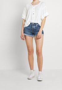 Bílá polo tričko s krátkým rukávem a knoflíky, spárované s modrými džínovými šortkami s roztrženými lemy. Oblečení doplňují bílé plátěné tenisky.