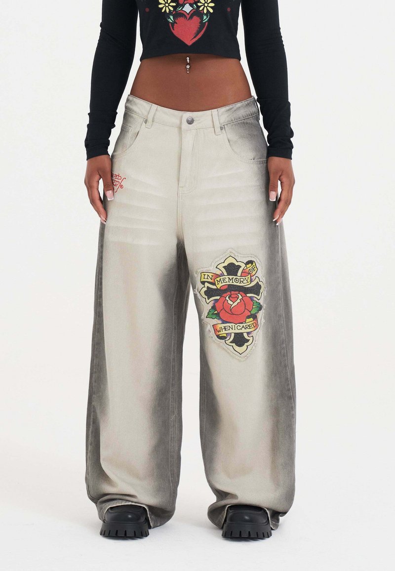Jeans de pierna ancha, de color gris desgastado, con un gran parche bordado de una rosa y el texto "En memoria de cuando me importaba" en el muslo izquierdo.