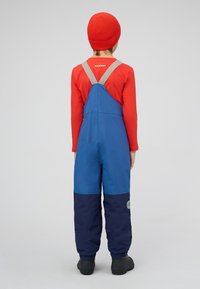 Blauw en marineblauw skipak met grijze bandjes, gewatteerde structuur en een logo-patch. Gecombineerd met een rood shirt met lange mouwen en een gebreide muts.