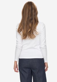 Weißes Langarmoberteil aus weichem Material, taillierter Schnitt, kombiniert mit dunklen Jeanshosen. Die Rückansicht zeigt eine schlichte Bauweise und eine glatte Textur.