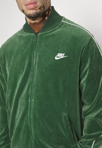 Chaqueta bomber Nike de terciopelo verde con rayas laterales blancas y logo, con cierre de cremallera y bolsillos laterales. Textura suave, diseño ajustado.