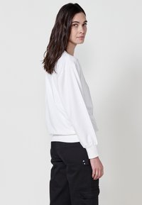 Felpa bianca dal fit rilassato, con spalle scese e polsini a costine. Abbinata a pantaloni cargo neri con tasche laterali e applicazioni metalliche.