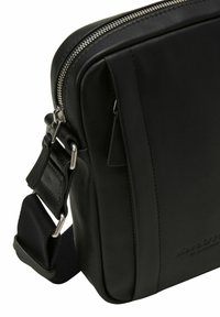 Sac à épaule en cuir noir avec une fermeture zippée, une sangle en tissu réglable et un détail de logo en relief. Texture lisse et forme rectangulaire.