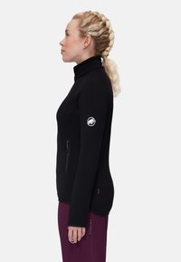 Giacca in pile nera con zip full-zip, colletto alto, design aderente, tasca con zip e patch logo sul braccio sinistro. Tessuto testurizzato.